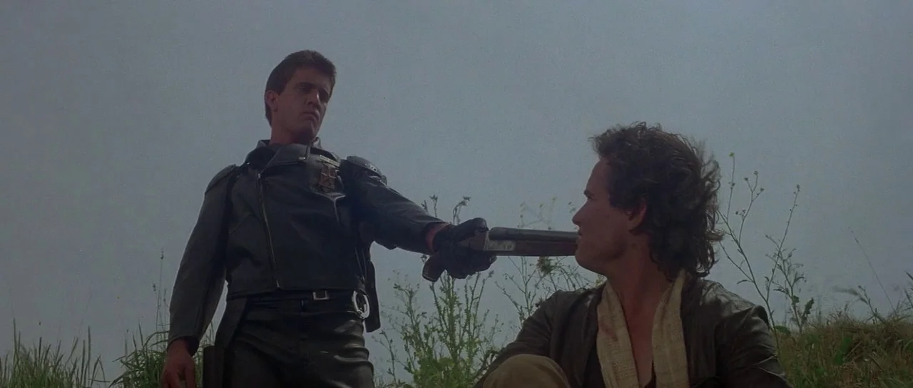 Mad Max (1979) movie still: medium — An open grassy field under a pale overcast sky at daylight. A man in black leather holds a shotgun against the…