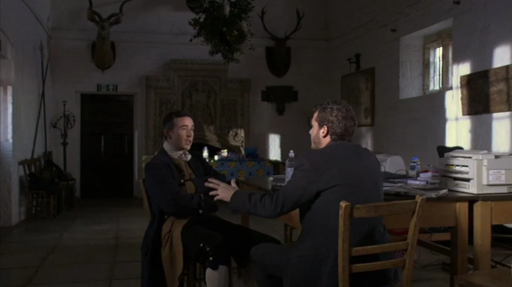 A Cock and Bull Story (2005) movie still: medium — A historic stone room features mounted animal heads on white walls and a large fireplace. Two men…