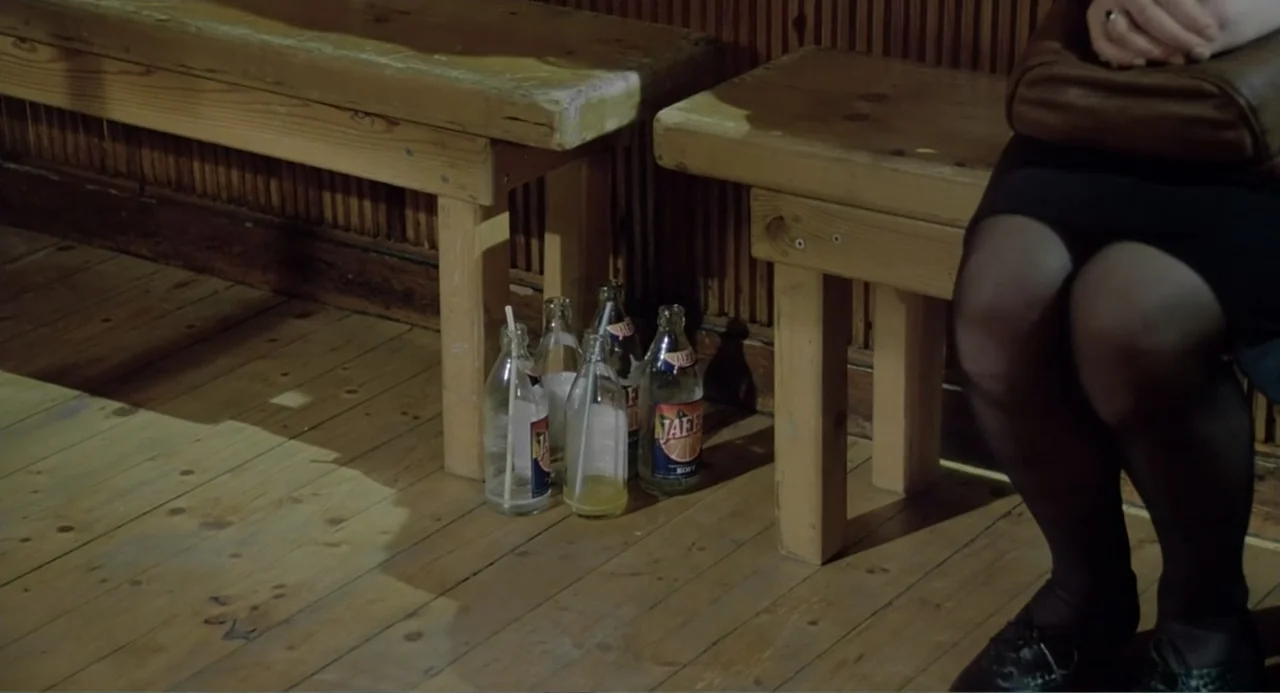 The Match Factory Girl (1990) movie still: medium — A wooden interior with benches and slatted walls in an older building. A person sits on the…