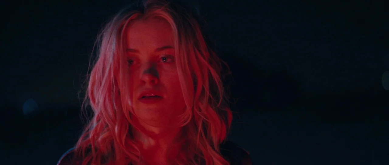 Starfish (2018) movie still: closeup — A woman stands in absolute darkness illuminated by a harsh red light source. The subject looks forward with…