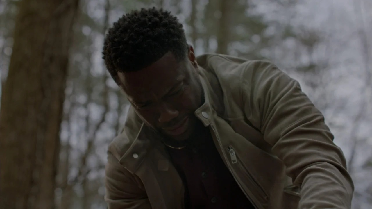 Die Hart (2023) movie still: medium — An outdoor forest setting features blurred tree trunks under an overcast grey sky. A man in a light jacket…