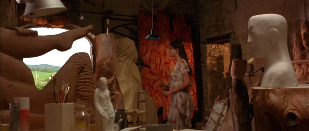 Stealing Beauty (1996) movie still: medium — A cluttered sculptor's studio contains numerous clay figures and wood carvings. A woman in a floral…