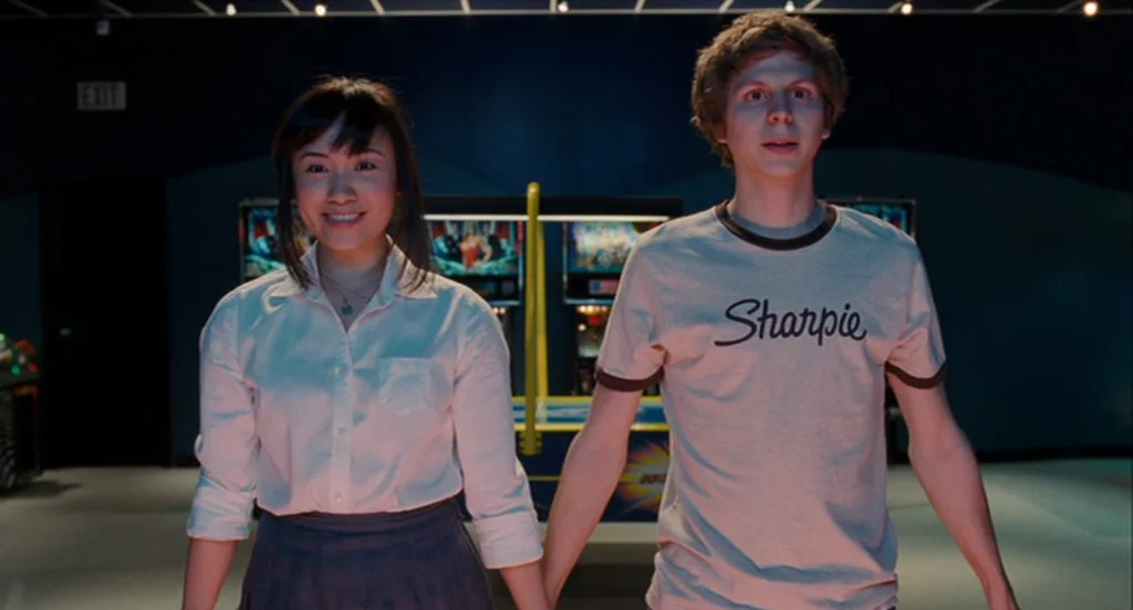 Scott Pilgrim Vs The World (2010) movie still: medium — A dimly lit arcade interior features blue walls with an illuminated exit sign. A young man…