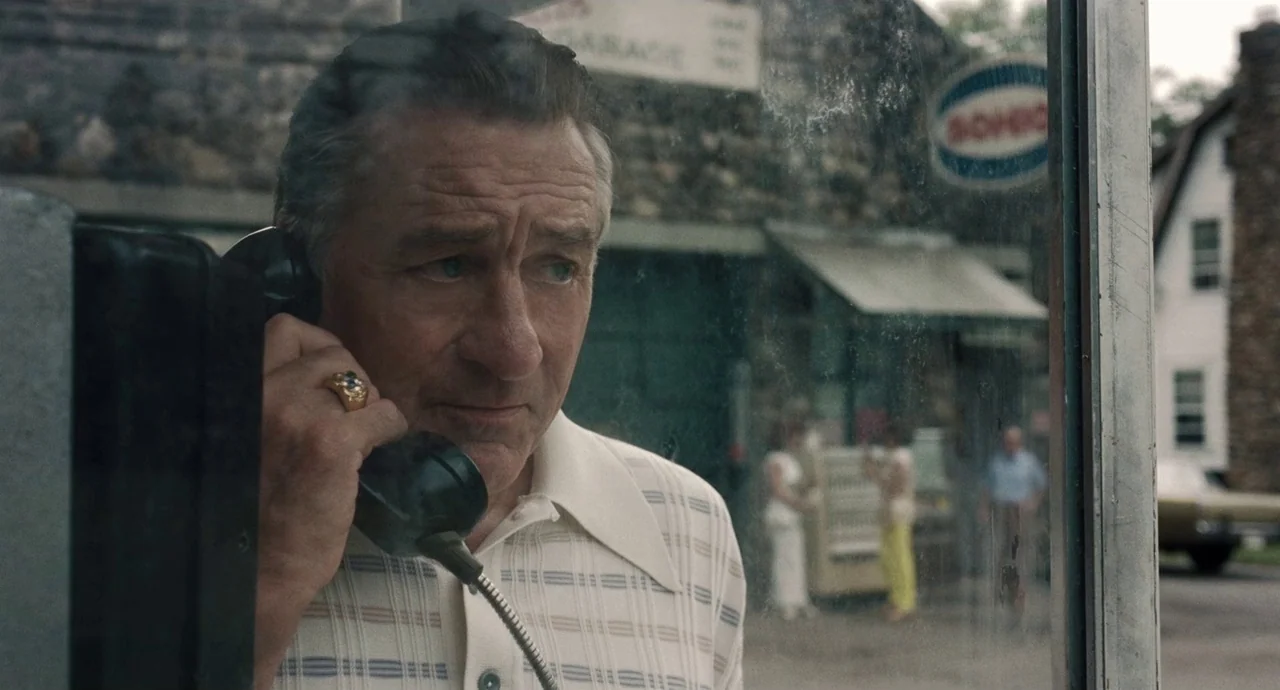 The Irishman (2019) movie still: closeup — An older man stands inside a glass phone booth outside a service garage. The man holds a telephone…