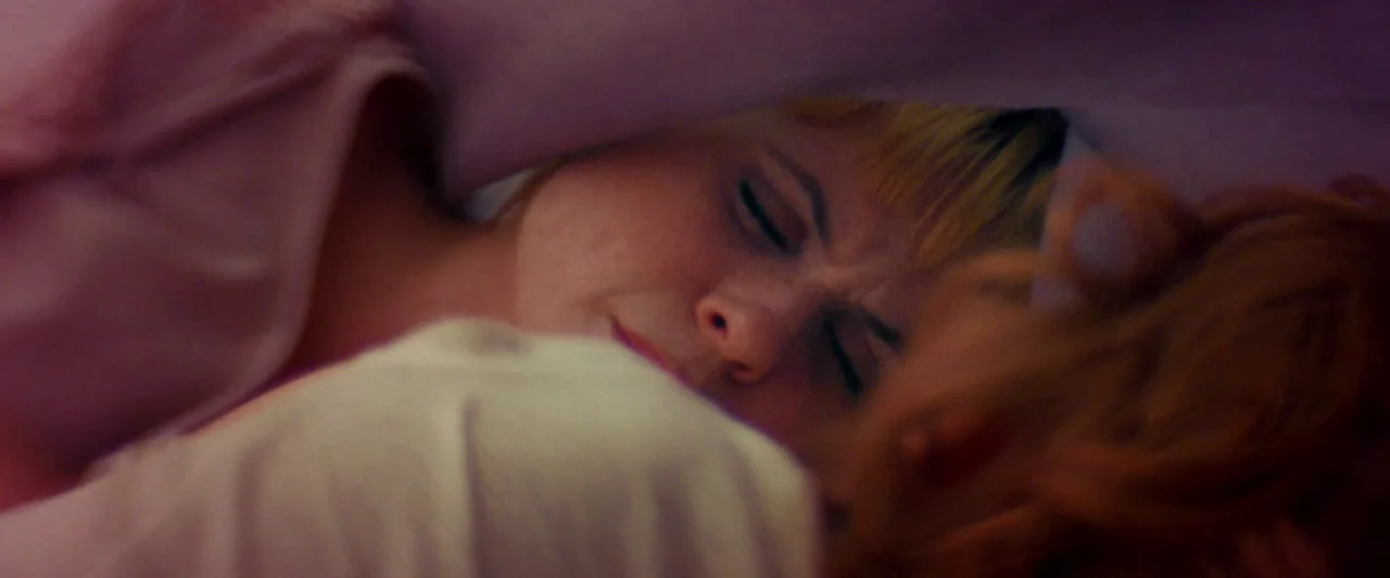 Moon Garden (2022) movie still: closeup — A person lies under a soft fabric blanket in a dim, purple-toned space. The individual rests with eyes…