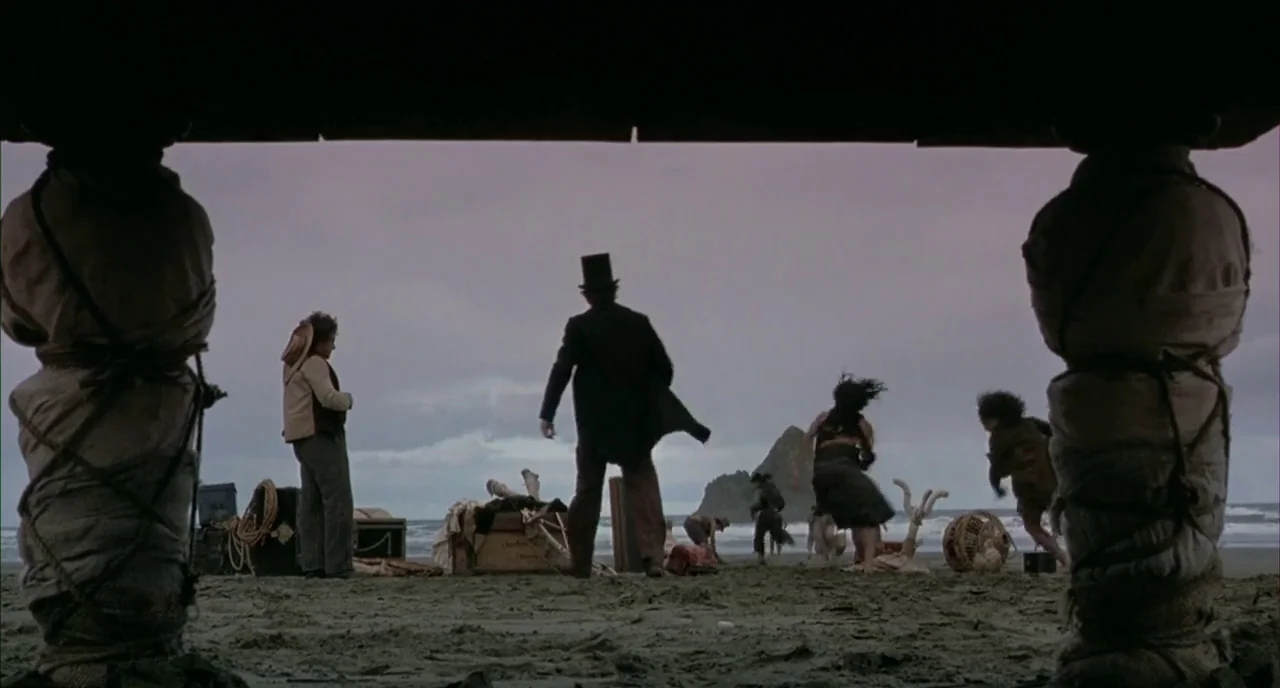 The Piano (1993) movie still: wide — A beach with dark sand under a cloudy sky near a large rock formation. Several people in 19th-century attire…