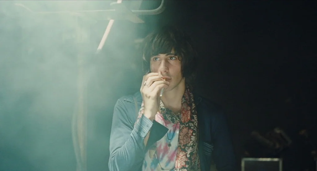Something in the Air (2012) movie still: medium — A dark space filled with light haze and mechanical equipment in the background. A young man stands…
