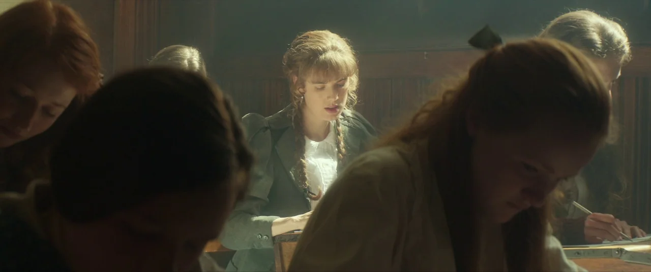 Sunset Song (2015) movie still: medium — A group of students sit at wooden desks in a sunlit academic space. A girl with braided hair concentrates…