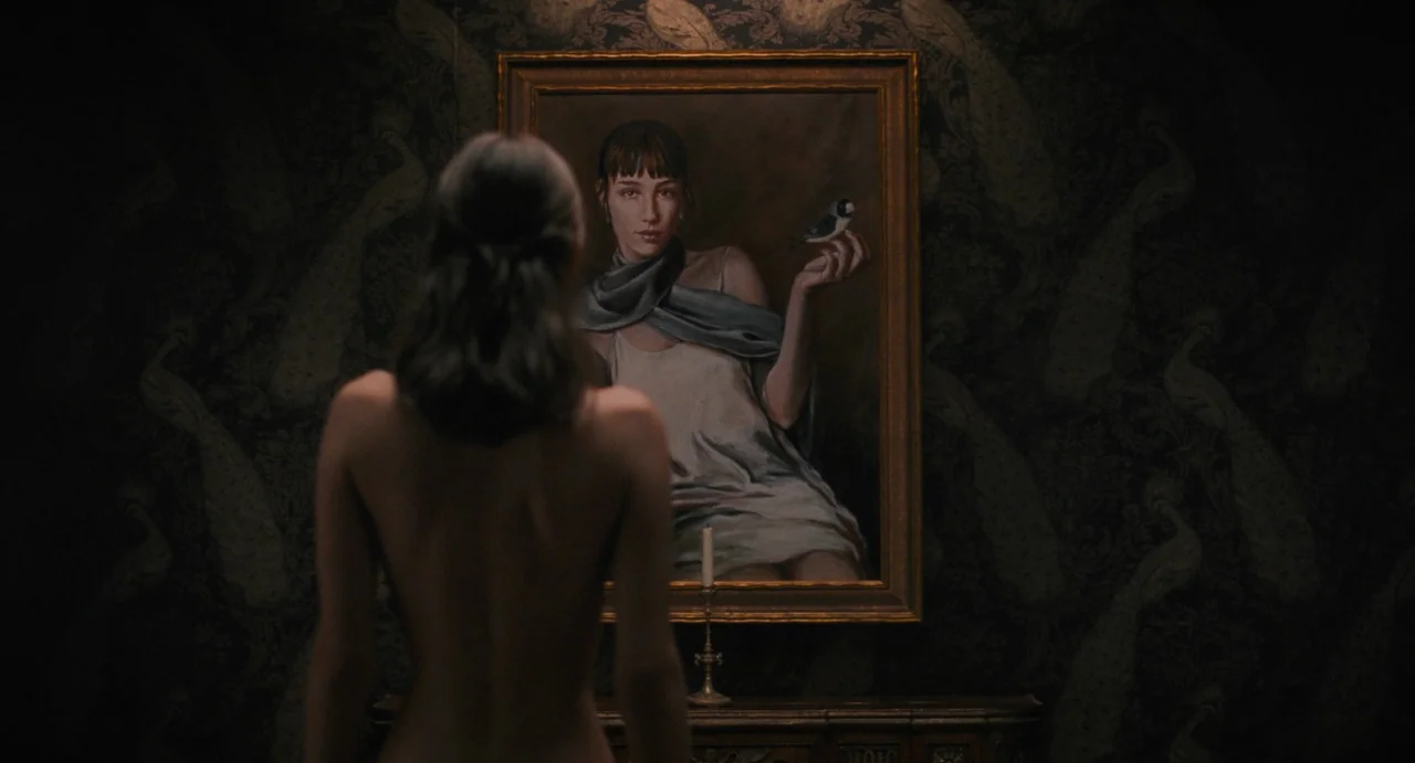 La Abuela (2021) movie still: medium — A dark study features black wallpaper with a subtle peacock feather pattern. A bare-shouldered woman stands…