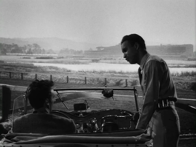 The Killing (1956) movie still: medium — A field at a racetrack with a large grandstand visible in the distance. One man stands by a convertible,…