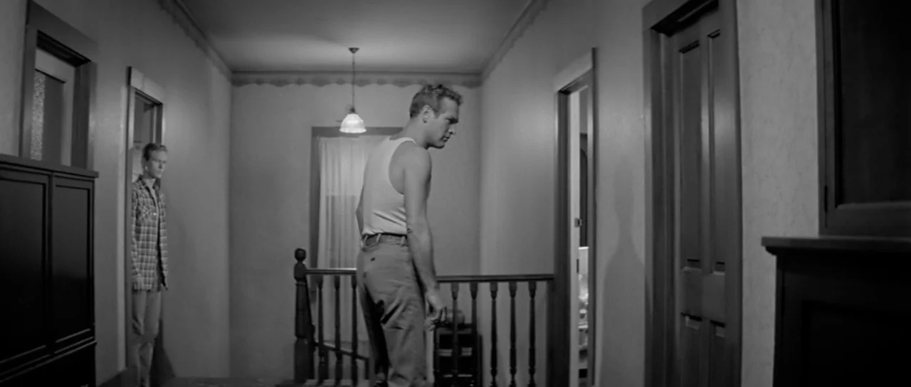 Hud (1963) movie still: medium — A dark, wood-trimmed residential hallway with multiple doorways and a staircase railing. A man in a tank top stands…