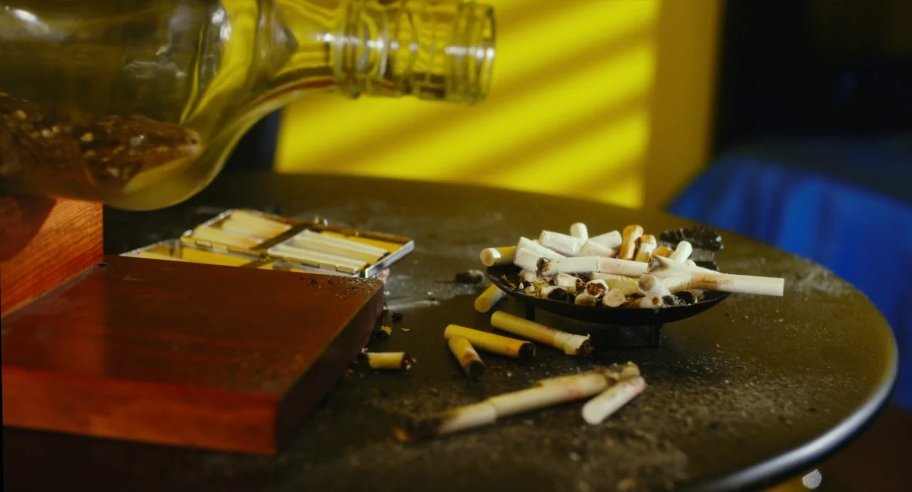 Making of: Antiporno (2016) movie still: closeup — A dark tabletop holds a crowded ashtray and a small metal cigarette case. Slatted light casts…