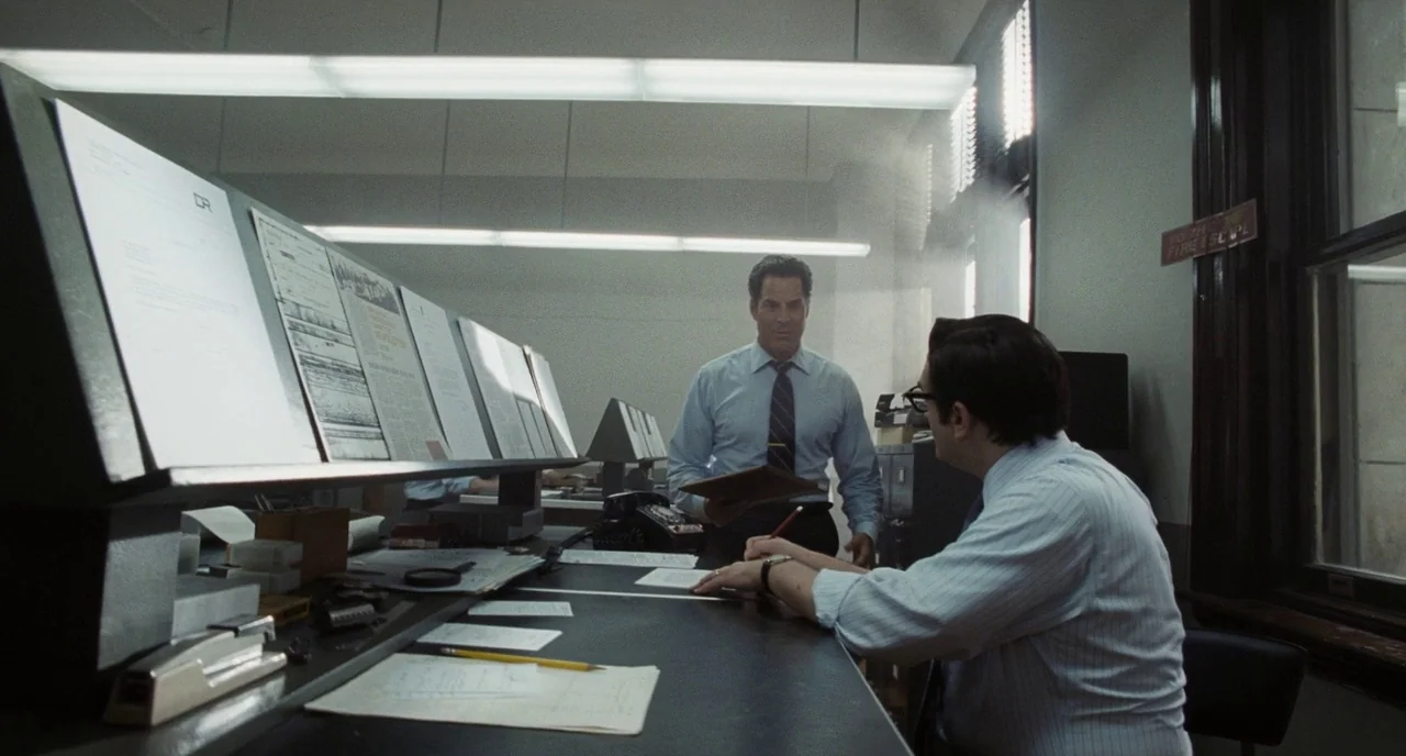 The Post (2017) movie still: medium — A 1970s newspaper office with angled document racks and bright fluorescent lighting. One man approaches his…