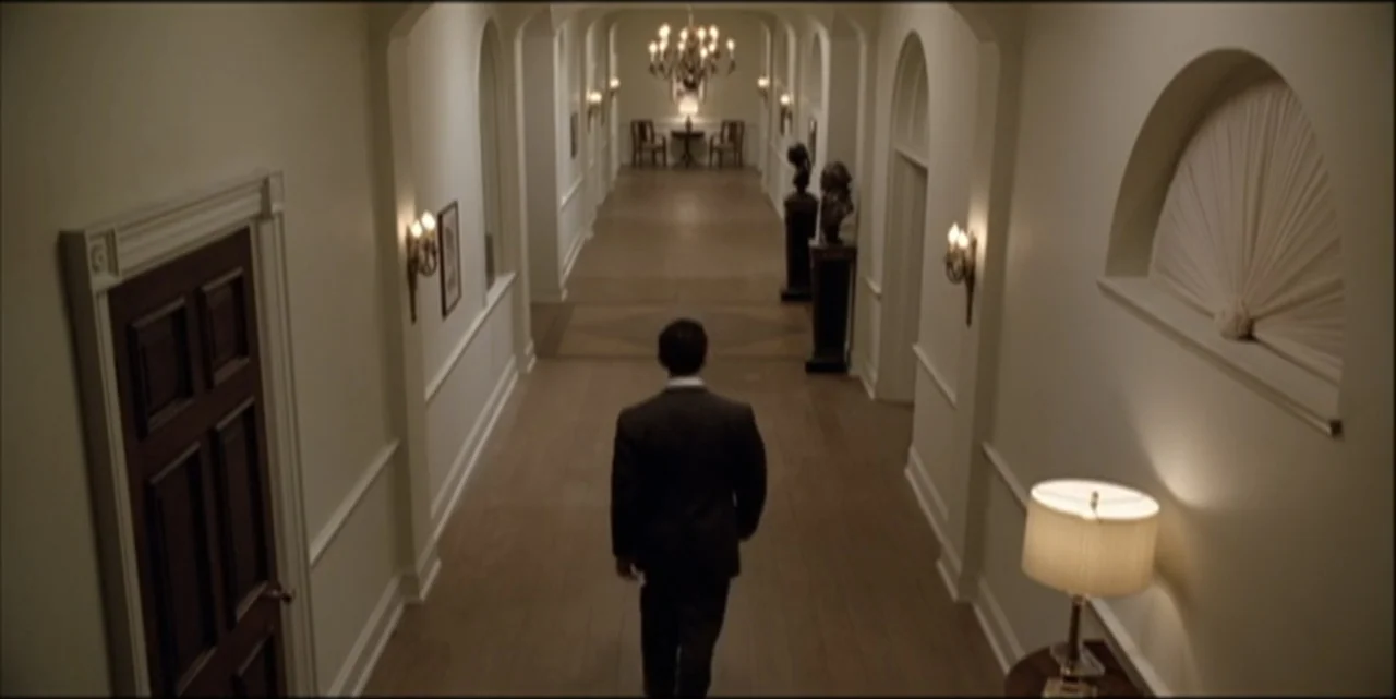 Path to War (2003) movie still: medium — A long, formal residential hallway features light-colored walls, wood floors, and arched alcoves. A man in…