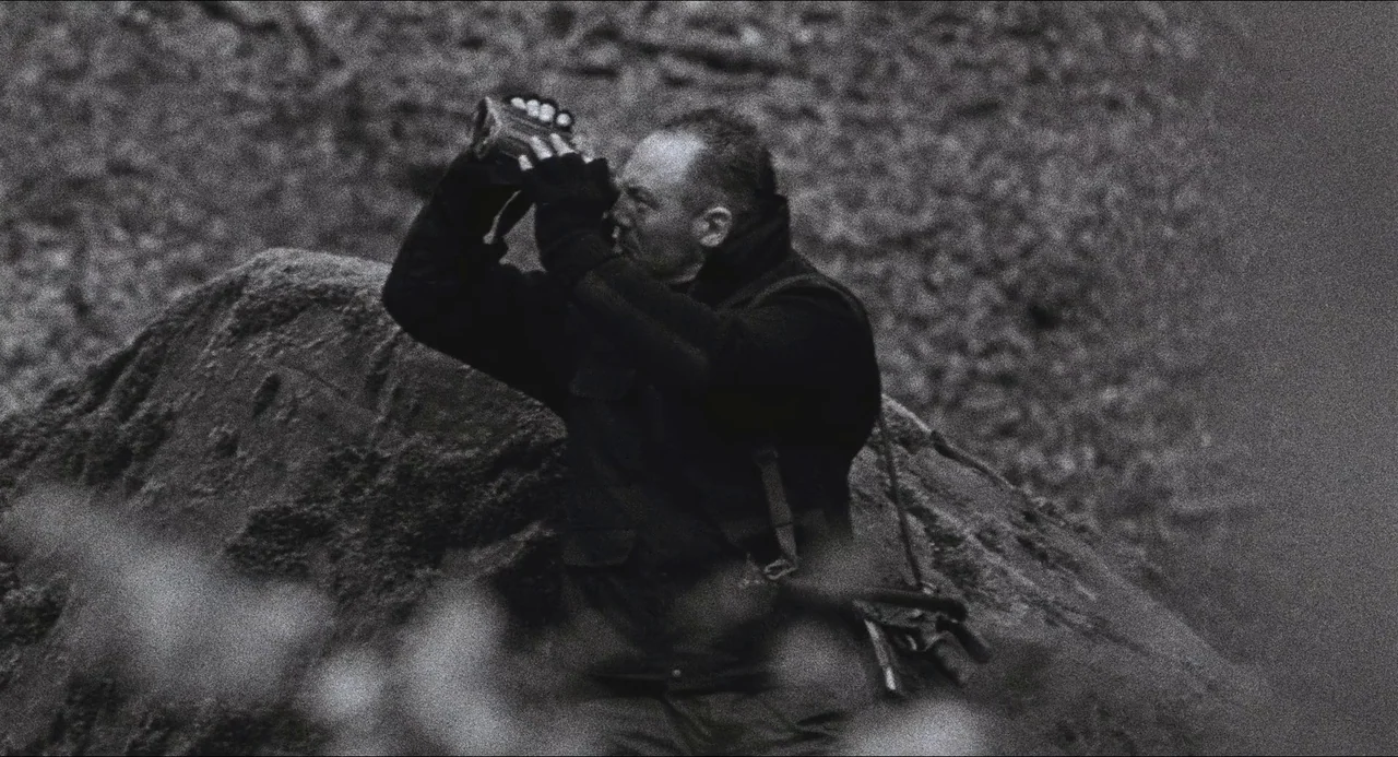 Dog Soldiers (2002) movie still: medium — A man crouches in a rugged, rocky outdoor environment with textured stone surfaces. He uses black…