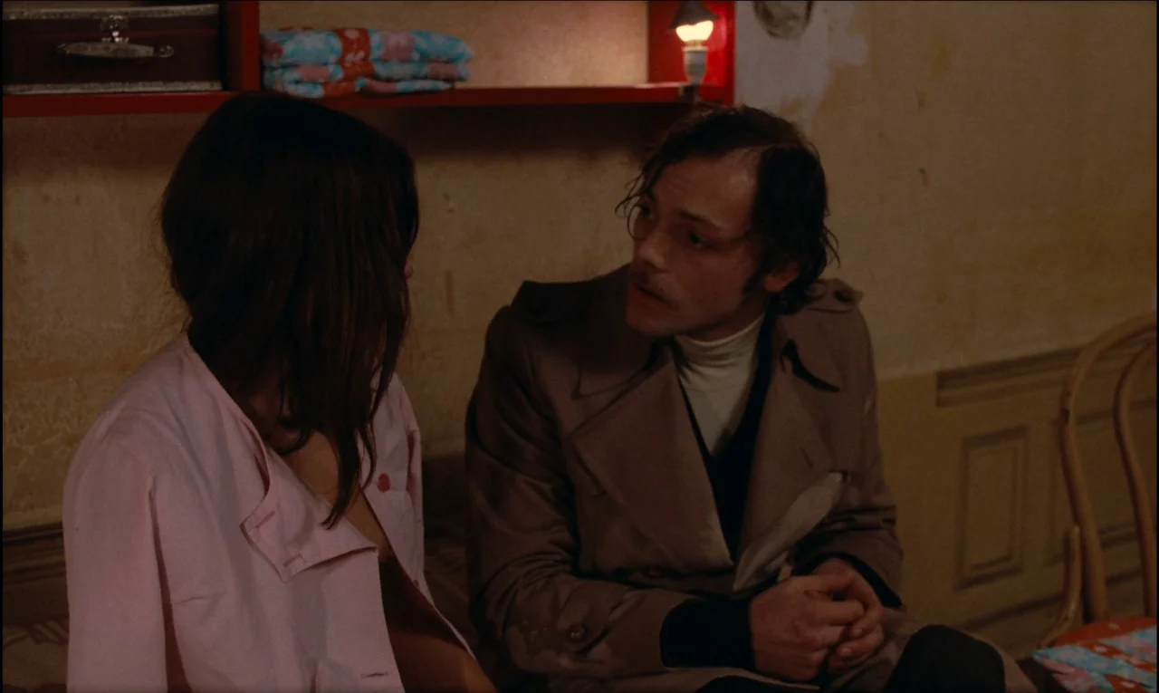 Serie Noire (1979) movie still: medium — A small indoor space with peeling beige walls and a red shelf holding folded linens. A woman in a pink…