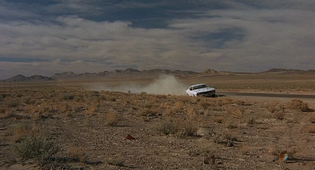 Vanishing Point (1971) movie still: wide — A vast, arid desert expanse features low rolling mountains against a cloudy blue sky. A solitary white…