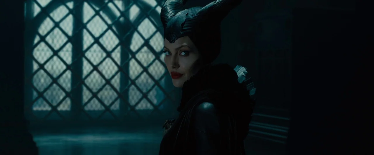 Maleficent (2014) movie still: medium — A dark stone room contains a large arched window with a metal lattice grid. A woman wearing large black…