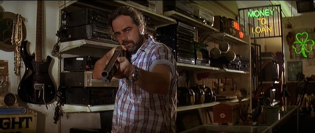 Pulp Fiction (1994) movie still: medium — A cluttered interior space filled with shelves of electronics and pawn shop goods. A bearded man in a…