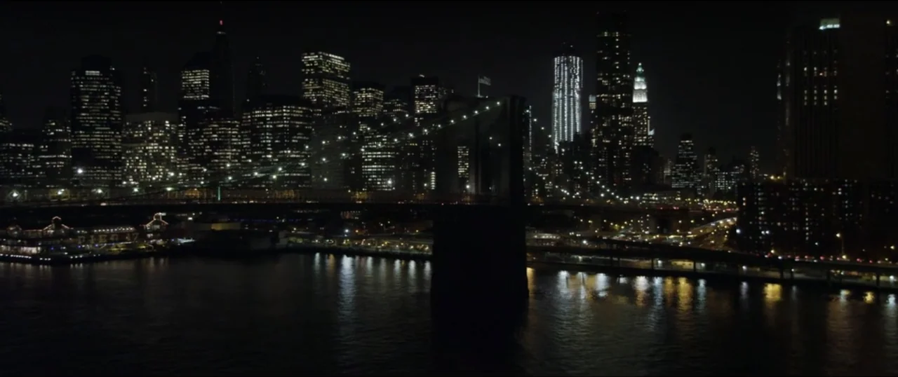 Scream VI (2023) movie still: establishing — The New York City skyline glows at night behind the dark silhouette of a bridge. Water reflects the…