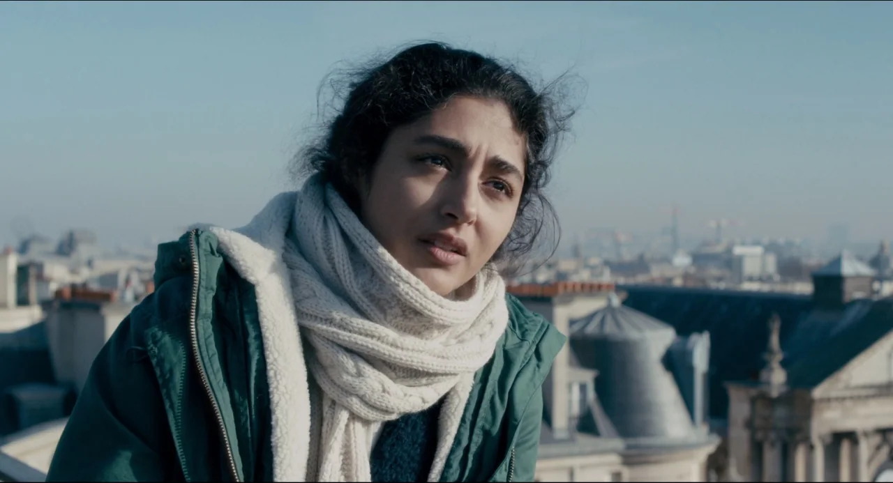 The Night Eats the World (2018) movie still: medium — A woman stands on a Parisian rooftop with classic zinc roofing visible below her. She looks…