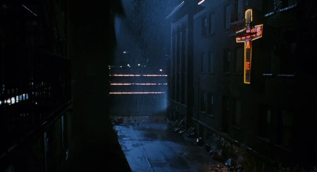 Mimic (1997) movie still: establishing — A rain-slicked dark alleyway is framed by brick buildings with visible fire escapes. An orange neon cross…