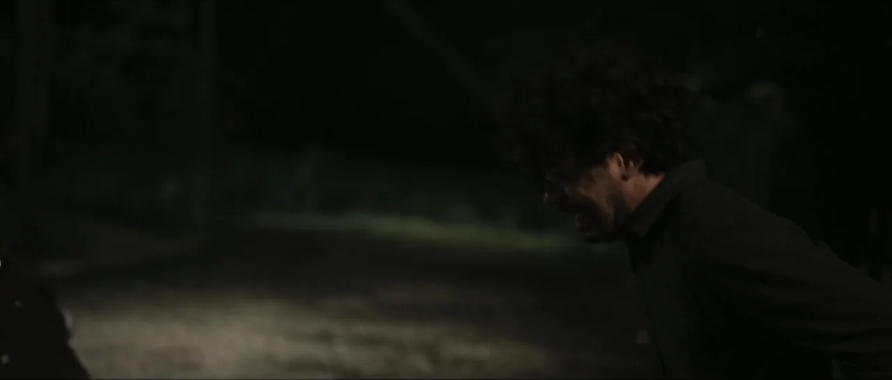 Innocent Vengeance (2023) movie still: medium — A dark outdoor space at night with a single lit area on the ground. A man with dark curly hair…