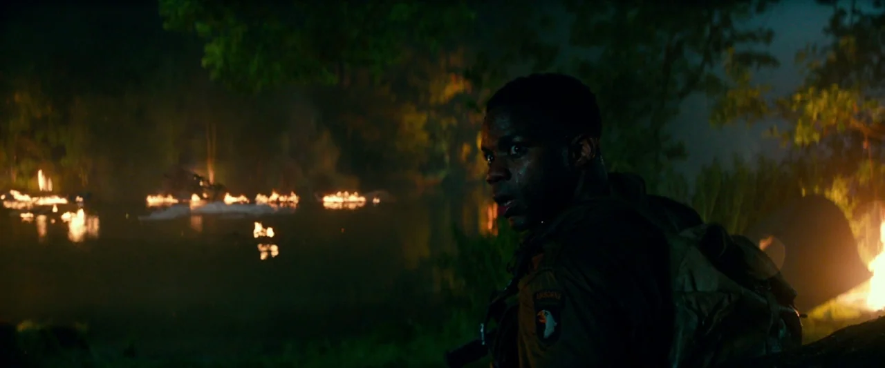 Overlord (2018) movie still: medium — A dense dark jungle at night with a small fire burning by the water. A soldier in uniform watches the flames…