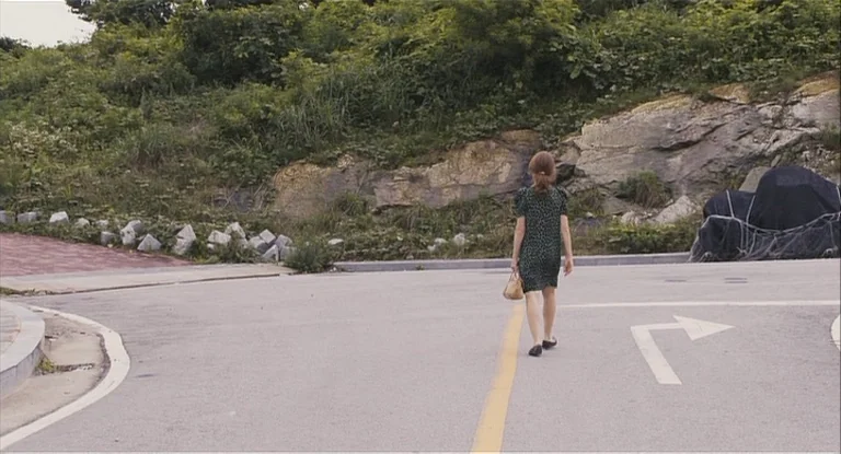 In Another Country (2012) movie still: fullbody — A paved intersection meets an embankment covered in dense green shrubs and rock outcroppings. A…