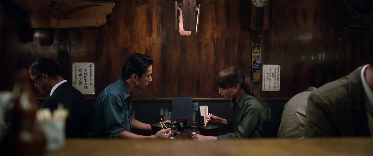 Earthquake Bird movie still: medium — A dimly lit Japanese noodle shop features dark wood paneling and aged wall posters. A man and a woman sit at…