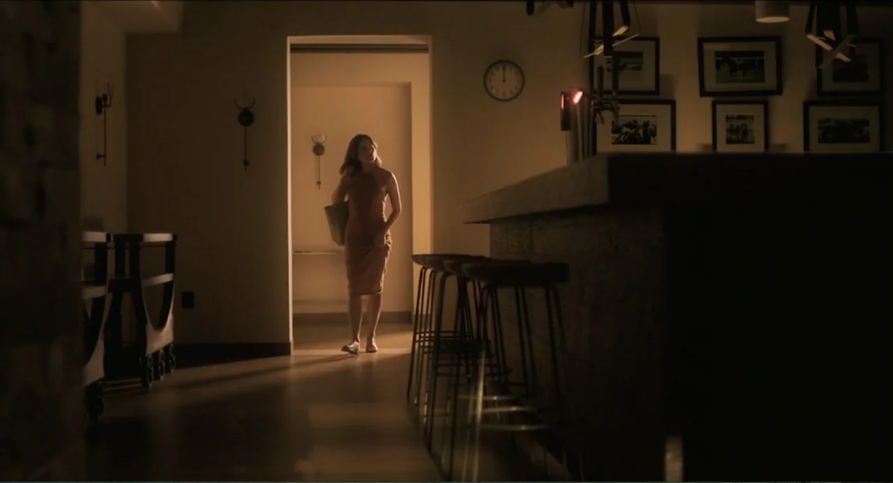At Midnight (2023) movie still: fullbody — A dimly lit hotel bar area features a counter, bar stools, and framed artwork on walls. A woman in a…