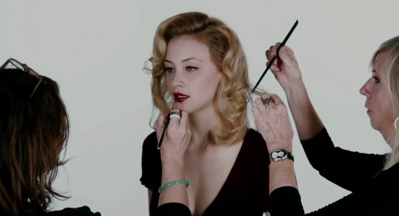 Antiviral (2012) movie still: medium — A woman sits in a brightly lit studio with a clean white backdrop. She is receiving hair styling and makeup…