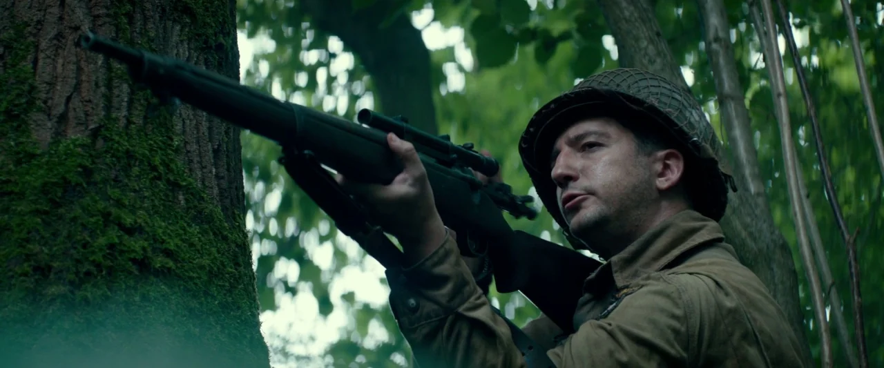 Overlord (2018) movie still: medium — A soldier in uniform stands among dense trees with moss-covered trunks. The man aims his rifle towards an…