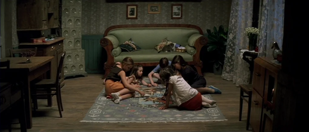 The Banishment (2007) movie still: wide — A furnished room features a green sofa, patterned area rug, and a tiled heater. Four children congregate…