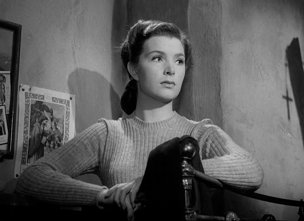 Odd Man Out (1947) movie still: medium — The wall behind the woman is plastered and decorated with religious images and a crucifix. The woman sits…