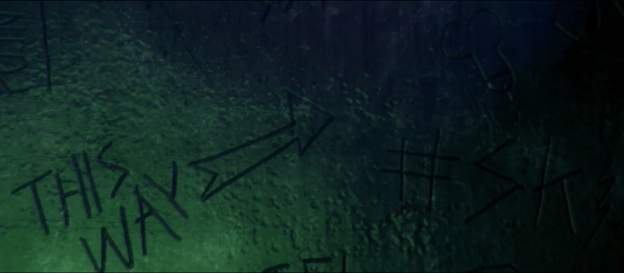 Guns Akimbo (2020) movie still: closeup — A dark, textured wall surface contains multiple hand-carved markings including an arrow pointing right.…