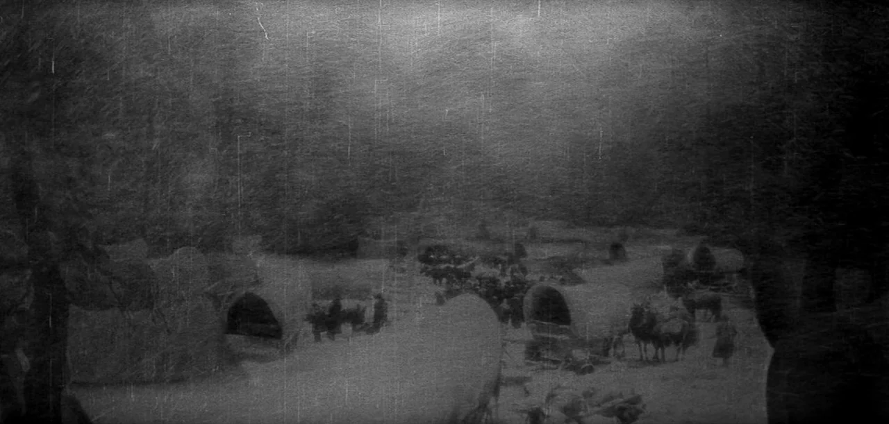 The Big Trail (1930) movie still: wide — A collection of canvas-covered wagons is dispersed across a dusty and barren dirt clearing. People stand…