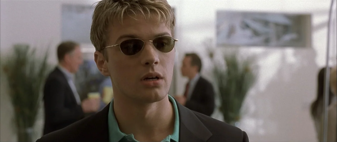 Igby Goes Down (2002) movie still: closeup — A brightly lit corporate building interior features a blurred background of office workers. A young man…