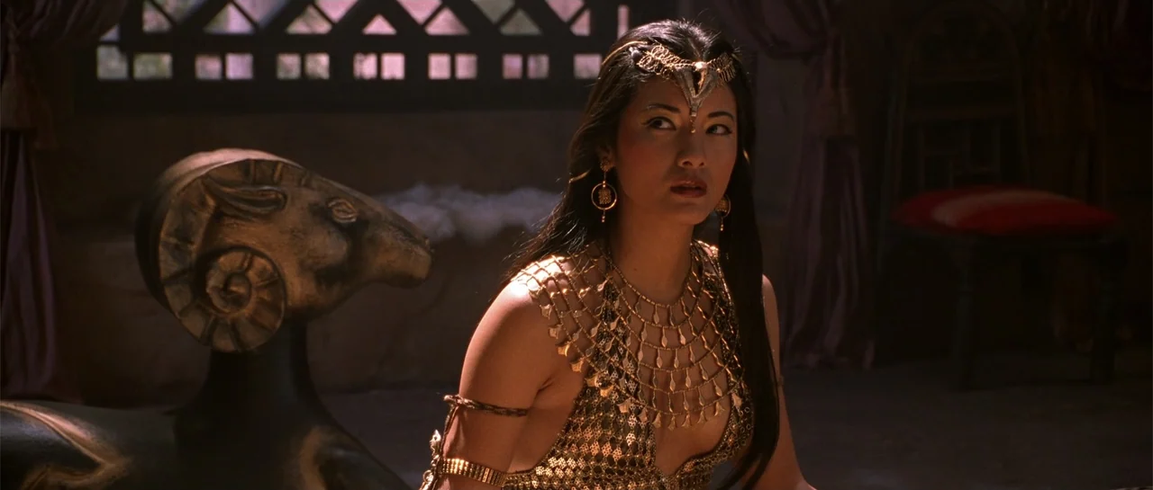 The Scorpion King (2002) movie still: medium — An interior chamber contains a bronze ram statue and textured stone walls. A woman wears golden armor…