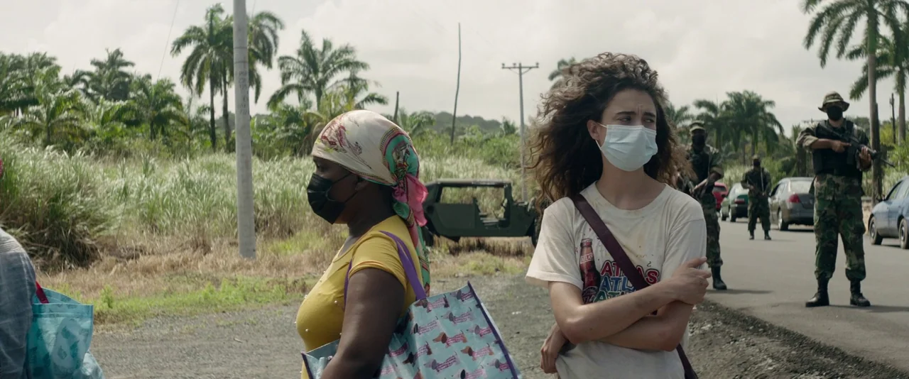Stars At Noon (2022) movie still: medium — A roadside scene features two women standing before a military checkpoint in a tropical area. Soldiers in…