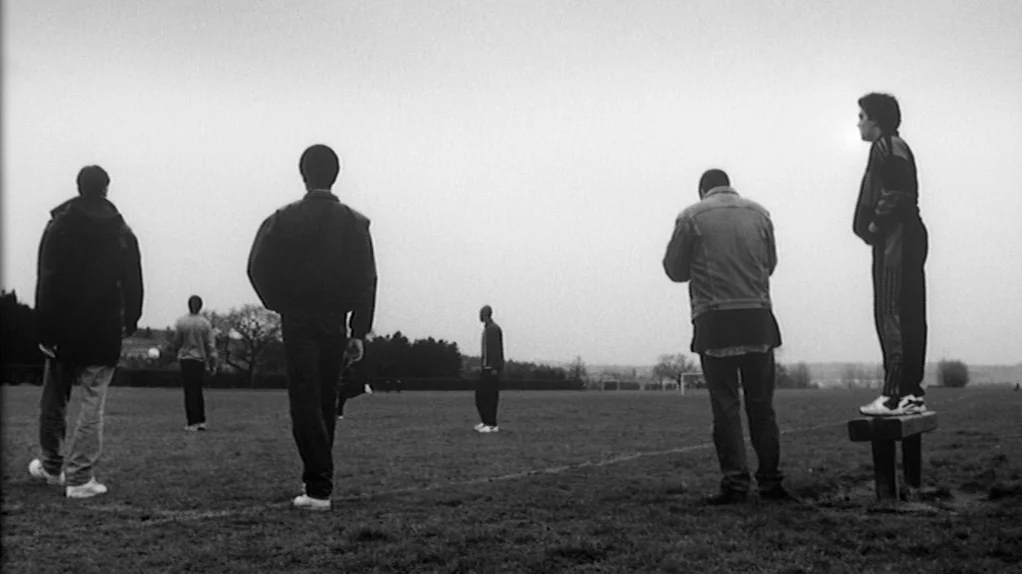 TwentyFourSeven (1997) movie still: wide — A vast, empty sports field under a gray, overcast sky with distant trees. Five young men stand at various…