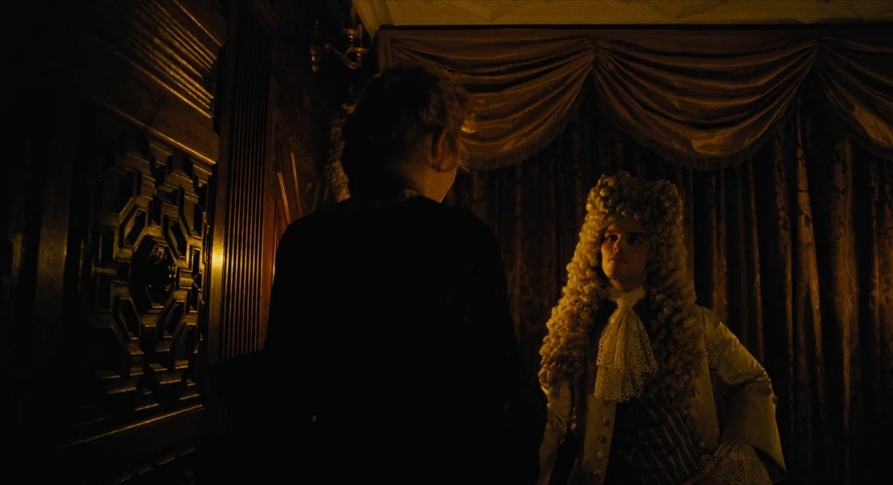 The Favourite (2018) movie still: medium — A dimly lit room with heavy brown velvet curtains and dark carved wooden paneling. A man in an…