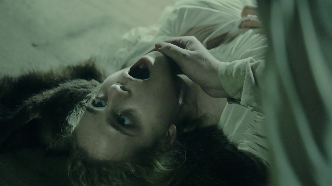 The Field Guide to Evil (2018) movie still: closeup — A woman lies on a wooden floor surface partially covered by a dark fur pelt. An unseen hand…