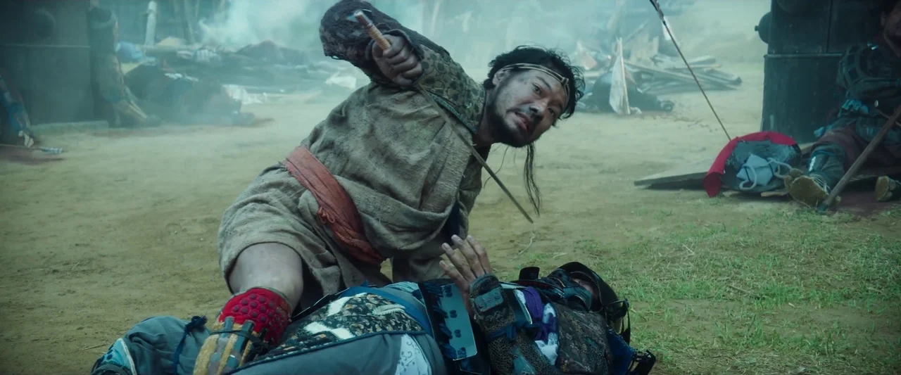 Kubi (2023) movie still: medium — A dusty, smoky battlefield littered with debris and scattered remnants of combat. A kneeling warrior holds a blade…