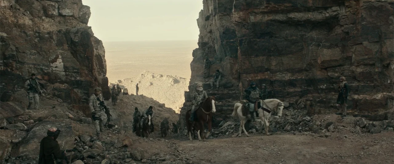 12 Strong (2018) movie still: wide — A rugged mountain pass features massive sheer rock walls and a narrow dirt path. Armed men in tactical gear and…