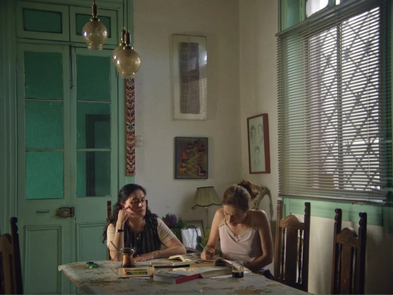 History and Geography (2024) movie still: medium — A room with pale walls, a floral tablecloth, and framed artwork hanging on the wall. Two women…