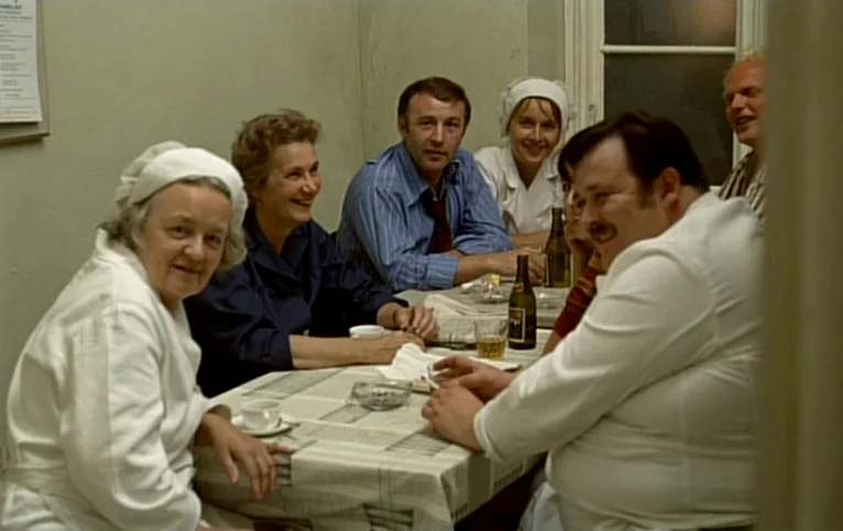 Giliap (1975) movie still: medium — A group of six people sit around a table with beverages and ashtrays. They appear to be engaged in conversation…