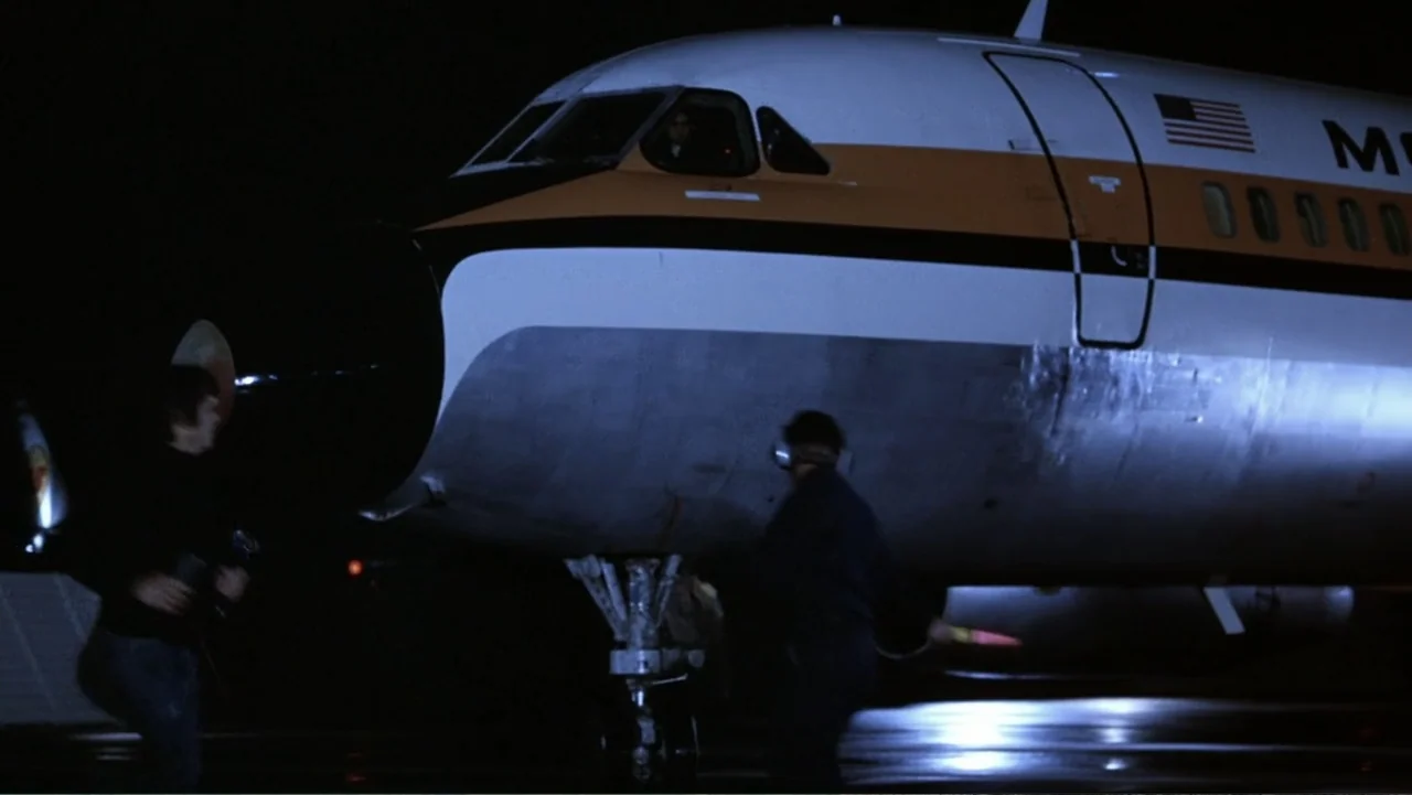 Dog Day Afternoon (1975) movie still: wide — A large commercial airplane sits on a dark airport tarmac at night. Two figures stand near the nose of…