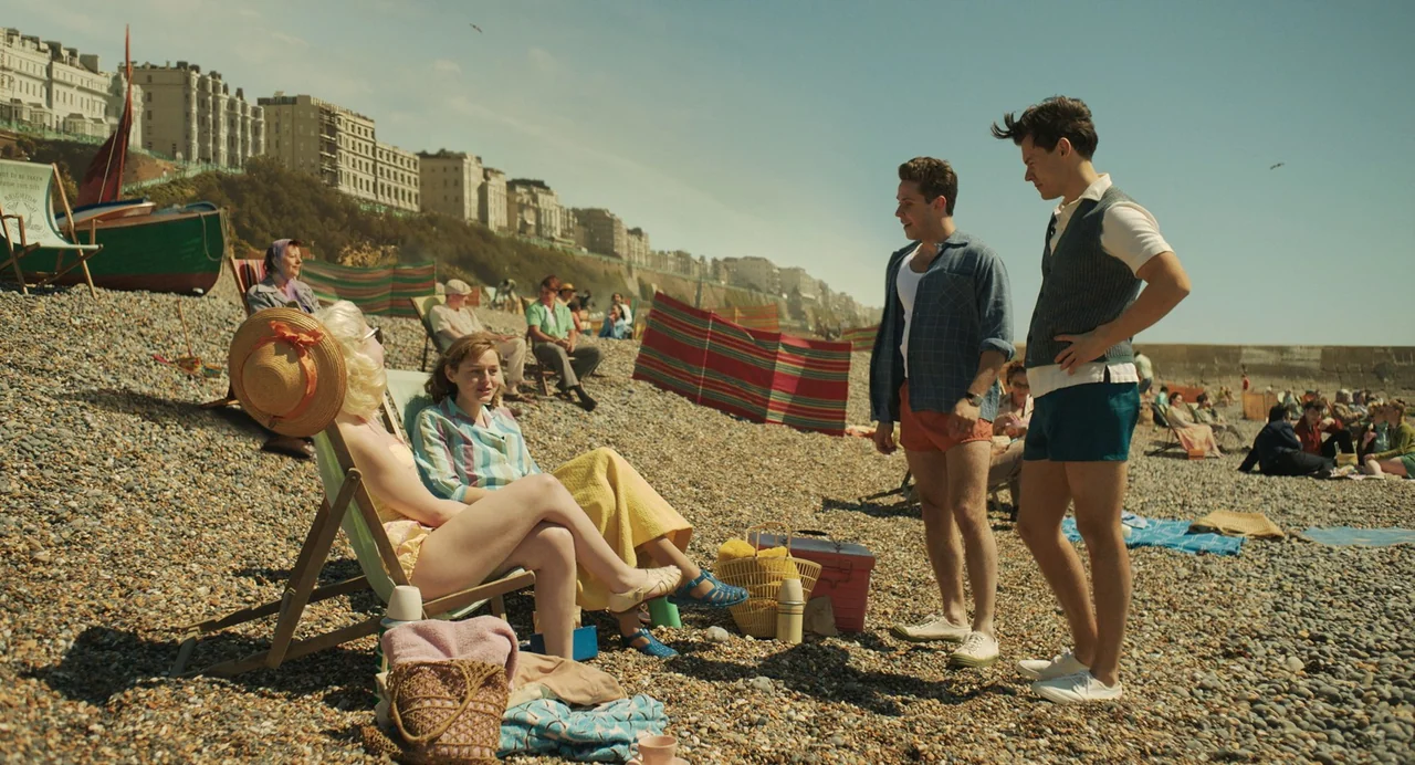 My Policeman (2022) movie still: wide — A pebble beach on a bright sunny day features rows of colorful windbreaks. Two men converse with two women…