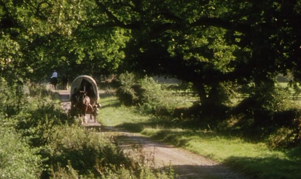 Black Jack (1979) movie still: wide — A rural dirt path winds through lush green foliage and large, mature oak trees. A horse-drawn covered wagon…